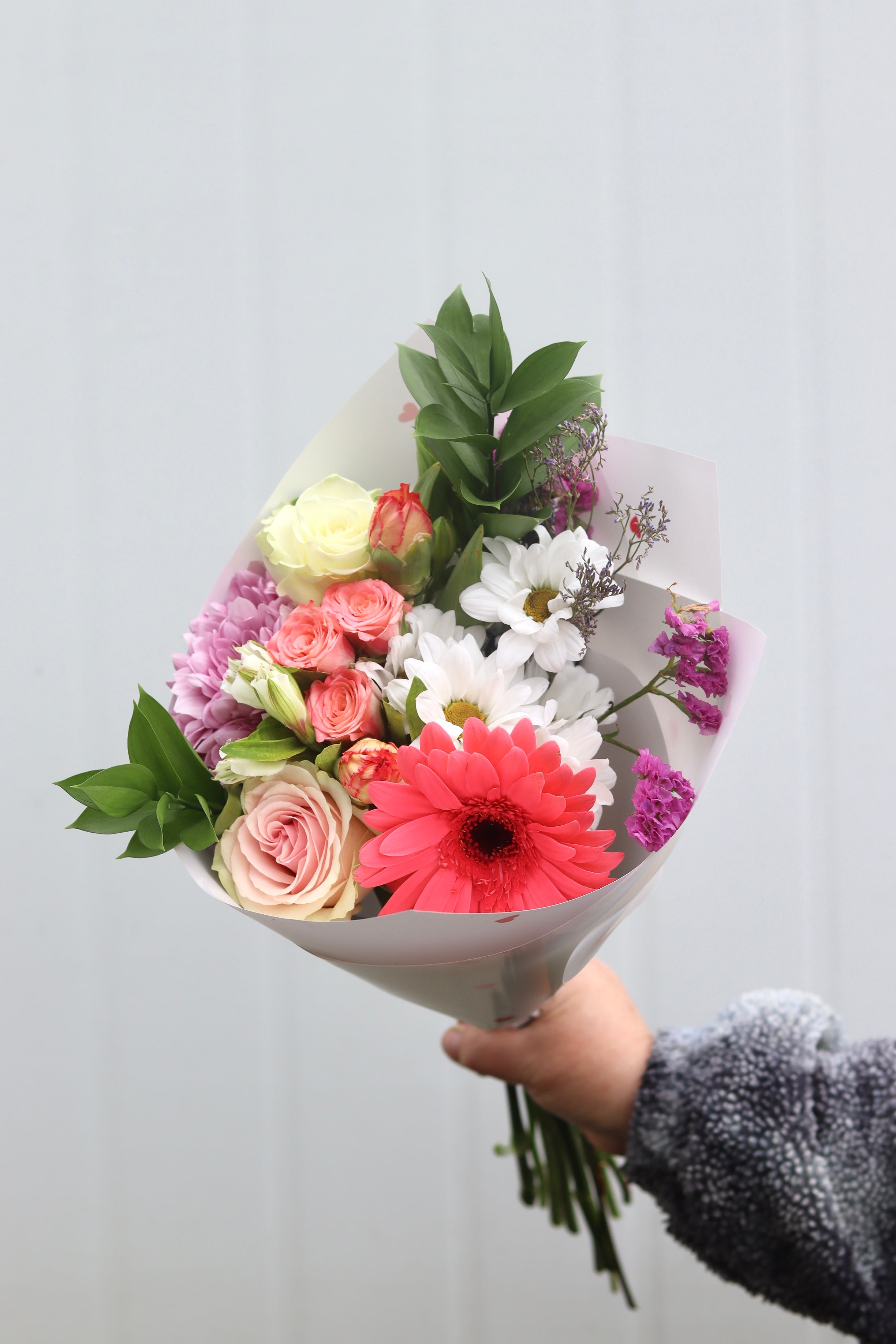 Ramo de flores tamaño S con gerbera fucsia, rosas en tonos crema y rosa, margaritas blancas, mini rosas coral y follaje verde, envuelto en papel blanco con acabado suave y sostenido por una mano.