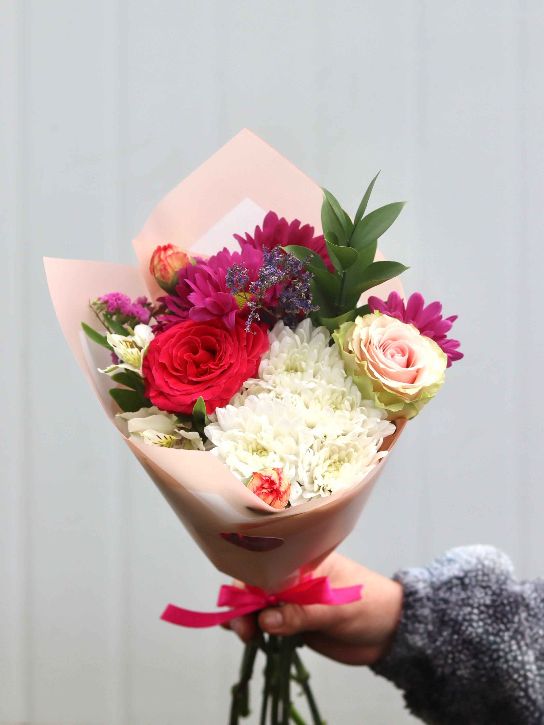 Ramo de flores tamaño XS envuelto en papel rosa claro, con una mezcla de flores frescas en tonos rosados, blancos y fucsias, que incluye rosas, crisantemos y follaje verde, sostenido por una mano y decorado con un lazo rosado.